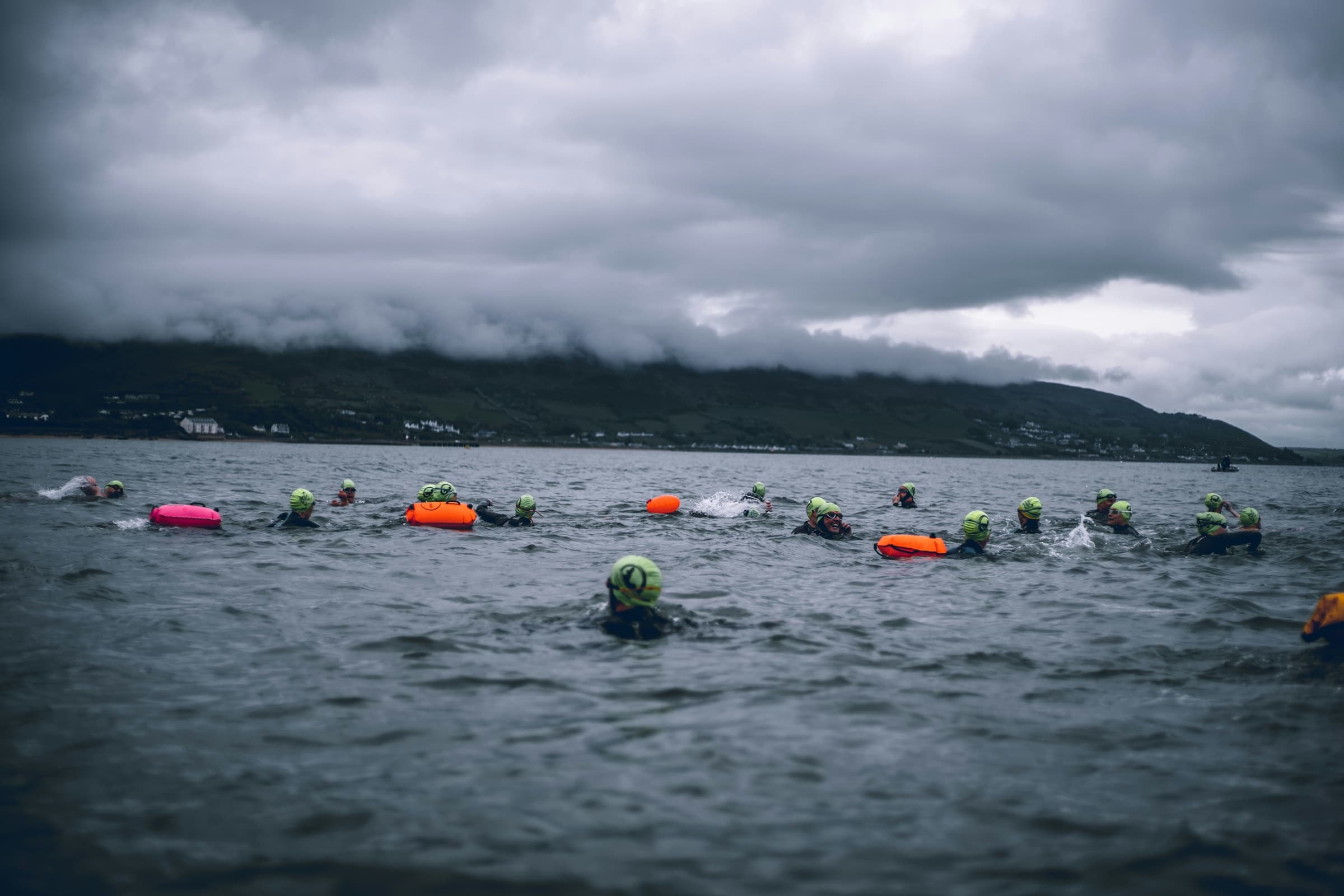 Open water swimmers wearing bright caps and tow floats beneath a stormy sky